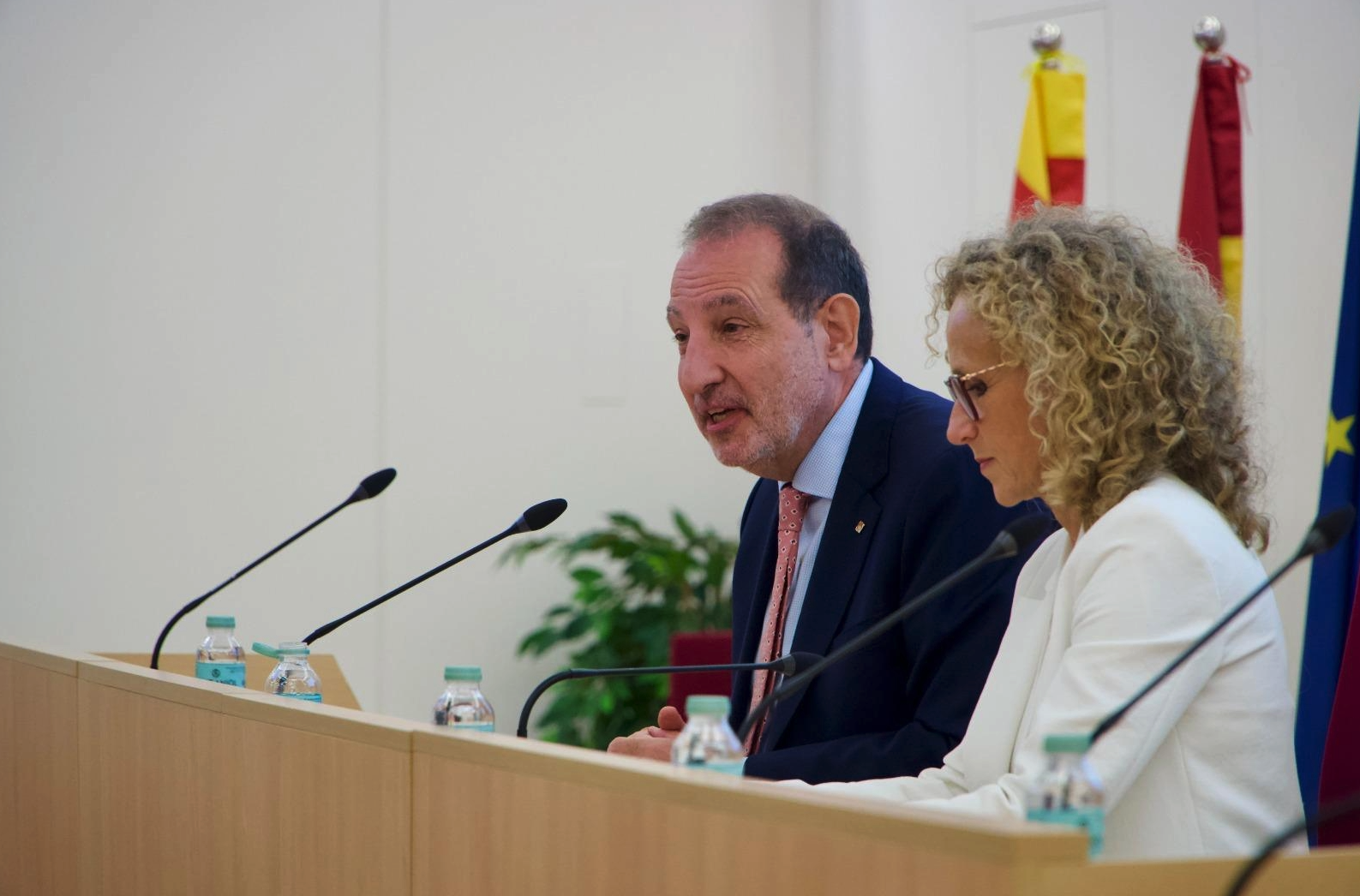 El director del Centre d’Estudis Jurídics i Formació Especialitzada, el Sr. Marc Cerón i Riera, intervé durant un acte institucional, assegut al costat de la secretària de Cicles de Vida i Ciutadania del Departament de Drets Socials i Inclusió, la Sra. Teresa Llorens Carbonell, davant d'una taula amb micròfons i amb les banderes institucionals al fons.